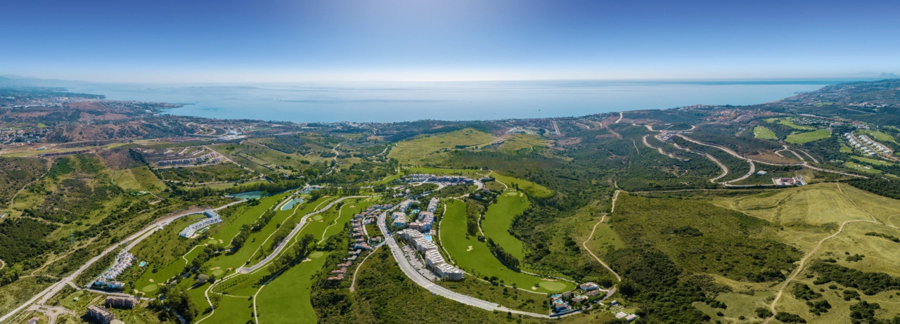 Una extensa vista aérea de la urbanización Sunny Golf, perfectamente integrada con las exuberantes calles verdes de Estepona Golf y el brillante mar Mediterráneo al fondo.